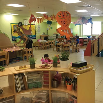 mosaic-nursery-reem-island- classroom-with-wooden-desks-and-chair-with-a-teacher-attending-to-kids