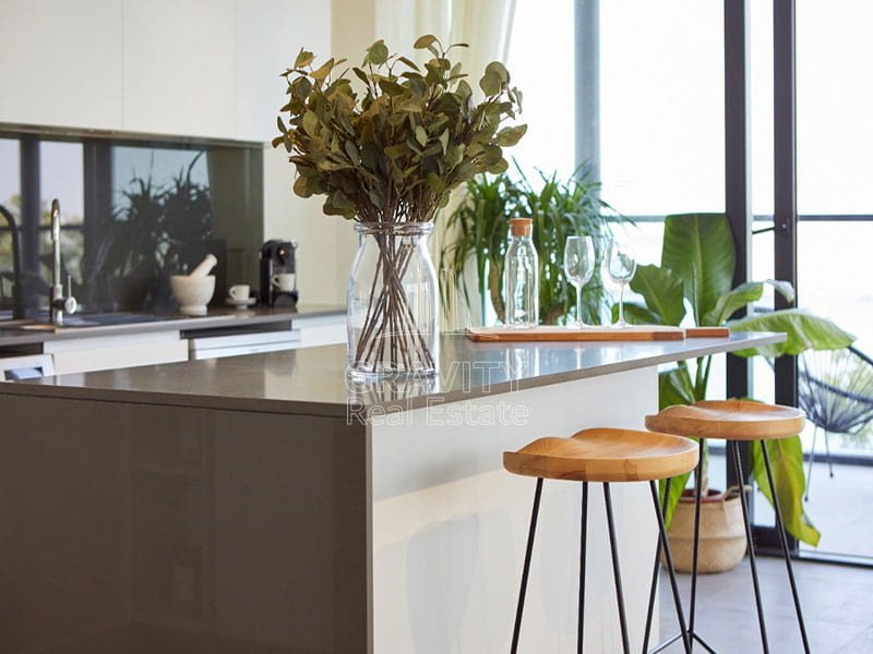 modern-kitchen-with-white-walls-and-light-wood-flooring. In-the-center-of-the-room-is-a-wooden-dining-table-surround-in-pixels